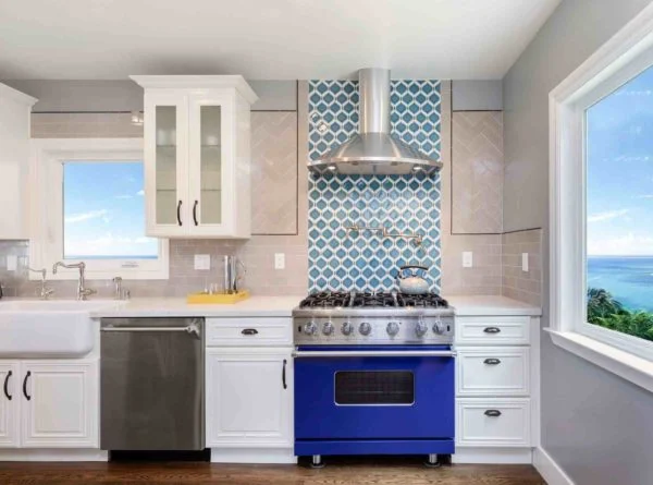 Bright kitchen with white cabinets, a blue oven, stainless steel appliances, and a blue patterned tile backsplash. A large window offers a scenic ocean view, reflecting popular 2021 Kitchen Design Trends. Bright kitchen with white cabinets, a blue oven, stainless steel appliances, and a blue patterned tile backsplash. A large window offers a scenic ocean view, reflecting popular 2021 Kitchen Design Trends.