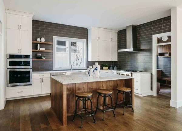 Modern kitchen with dark subway tile backsplash, white cabinets, a large wooden island with three stools, built-in ovens, and hardwood floors. Natural light enters through a window above the sink, reflecting current kitchen design trends. Modern kitchen with dark subway tile backsplash, white cabinets, a large wooden island with three stools, built-in ovens, and hardwood floors. Natural light enters through a window above the sink, reflecting current kitchen design trends.
