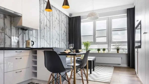 Modern kitchen and dining area featuring 2021 kitchen design trends—white cabinets, black chairs around a small round table, pendant lights, large windows with gray curtains, and potted plants on the windowsill. Modern kitchen and dining area featuring 2021 kitchen design trends—white cabinets, black chairs around a small round table, pendant lights, large windows with gray curtains, and potted plants on the windowsill.