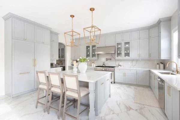 Modern kitchen design with light gray cabinetry, marble countertops and floors, gold pendant lights over an island with three upholstered stools, stainless steel appliances, and bright natural lighting. A showcase of current kitchen trends. Modern kitchen design with light gray cabinetry, marble countertops and floors, gold pendant lights over an island with three upholstered stools, stainless steel appliances, and bright natural lighting. A showcase of current kitchen trends.
