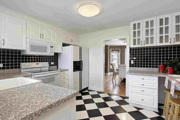 Modern kitchen design with white cabinets, granite countertops, black tile backsplash, and a black-and-white checkered floor. A microwave and refrigerator are visible, with a dining area seen through the doorway in the background—reflecting 2021 kitchen trends. Modern kitchen design with white cabinets, granite countertops, black tile backsplash, and a black-and-white checkered floor. A microwave and refrigerator are visible, with a dining area seen through the doorway in the background—reflecting 2021 kitchen trends.