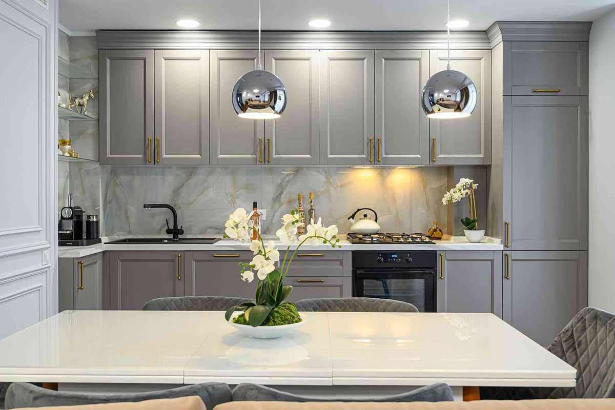 Modern kitchen with gray cabinets, gold handles, marble backsplash, built-in appliances, under-cabinet lighting, and two metallic pendant lights—showcasing top 2021 Kitchen Design Trends. White dining table with a floral arrangement in the foreground.