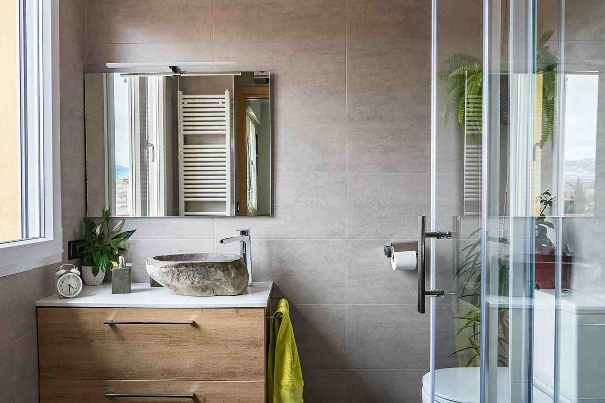 Modern bathroom design with a wooden vanity, stone sink, mirror, potted plants, and glass shower enclosure. Clever layout helps make the small bathroom bigger, while a yellow towel and natural light add warmth.