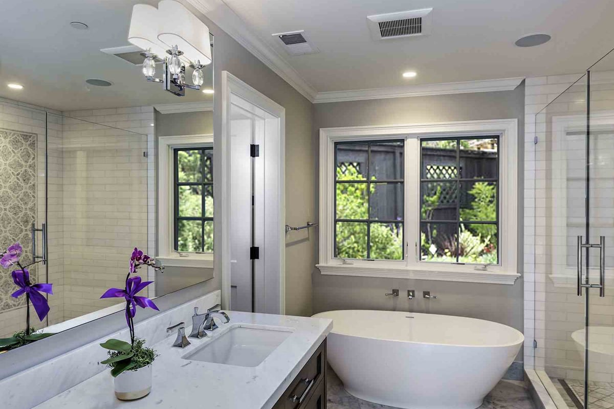 Modern master bathroom with a white freestanding tub under two windows, glass-enclosed shower, double sink vanity with orchid plants, and neutral-colored walls. Natural light enhances this stunning bathroom renovation.