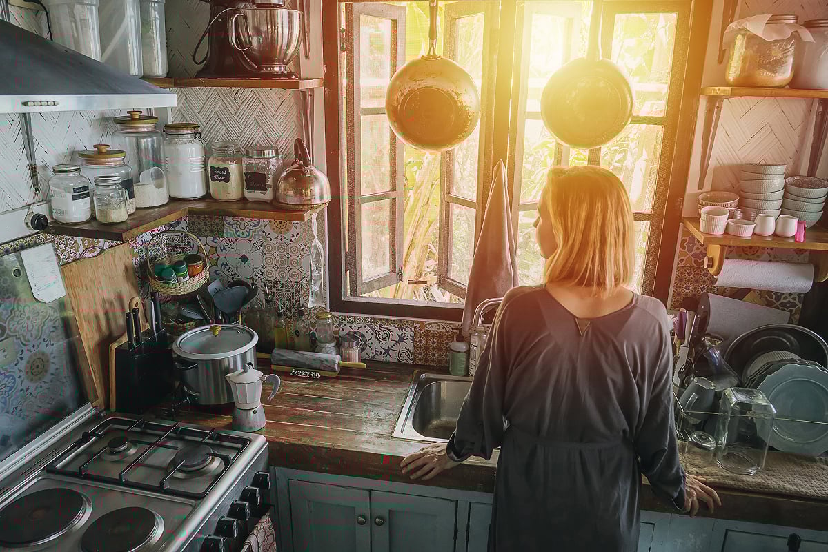 A woman stands at a kitchen sink, looking out an open window with sunlight streaming in. The kitchen remodel features shelves with jars, dishes, and cookware, plus patterned tiles on the walls.