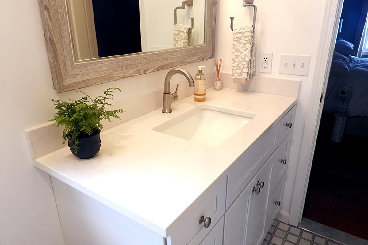 A bathroom vanity with a white countertop and built-in sink is perfect for any bathroom renovation, featuring a modern faucet, small potted fern, soap dispenser, reed diffuser, folded hand towel, and a large framed mirror above.