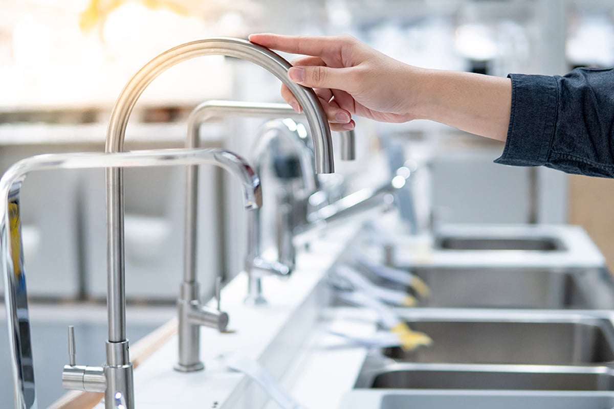 A person’s hand tests a modern, stainless steel kitchen faucet in a showroom, exploring different types of kitchen faucets displayed alongside various sinks in the background.