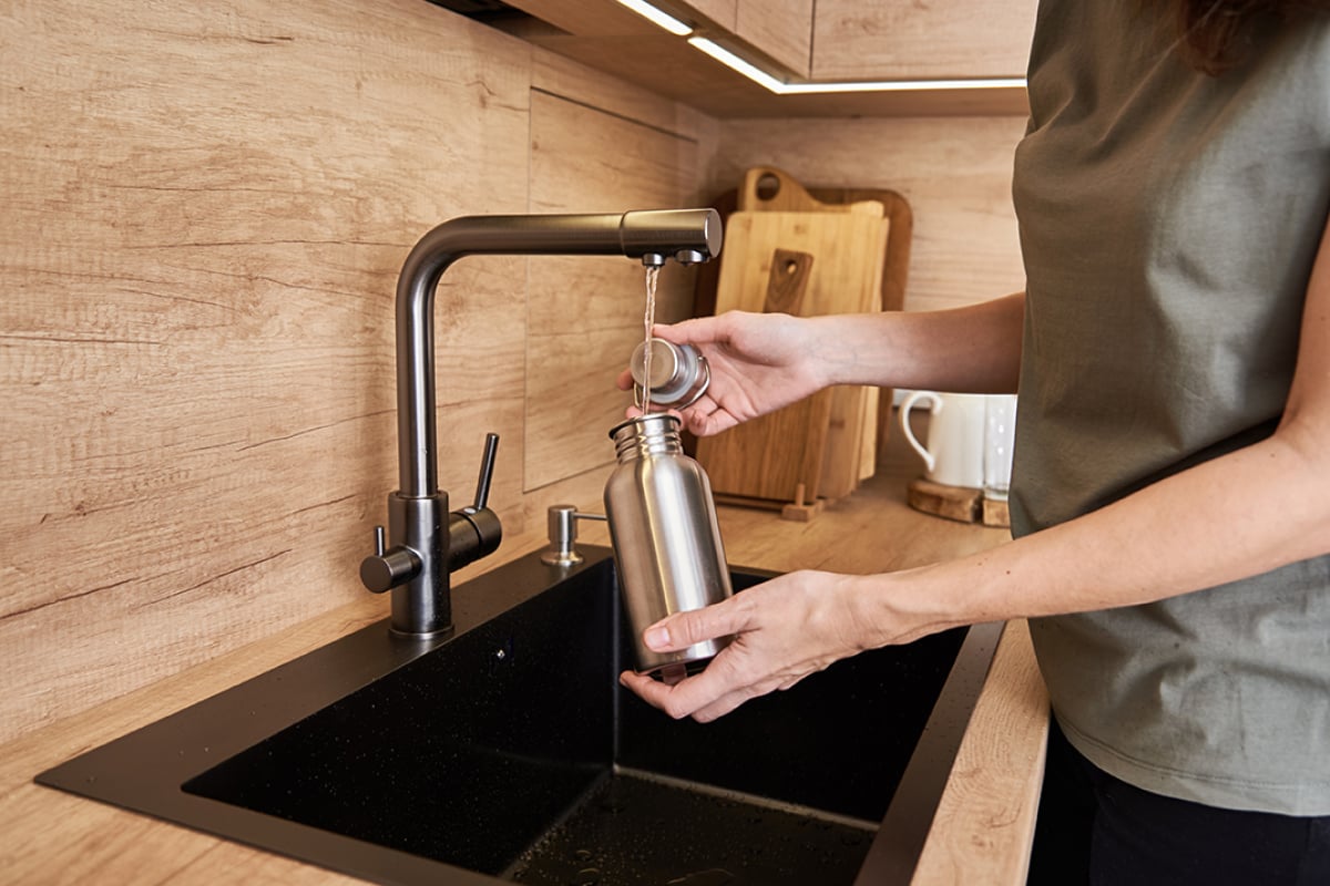 A person fills a stainless steel water bottle with water from one of the best kitchen faucets over a black sink, with wooden cabinets and cutting boards in the background. A person fills a stainless steel water bottle with water from one of the best kitchen faucets over a black sink, with wooden cabinets and cutting boards in the background.