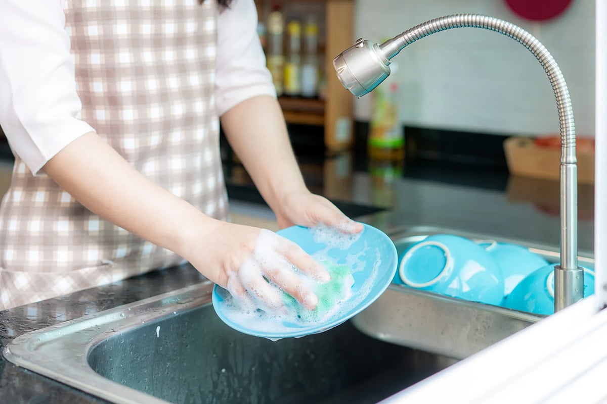 Person wearing a checkered apron washes a blue plate with soap and sponge at the kitchen sink, beneath one of the best kitchen faucets, with several clean blue bowls neatly arranged beside the sink. Person wearing a checkered apron washes a blue plate with soap and sponge at the kitchen sink, beneath one of the best kitchen faucets, with several clean blue bowls neatly arranged beside the sink.