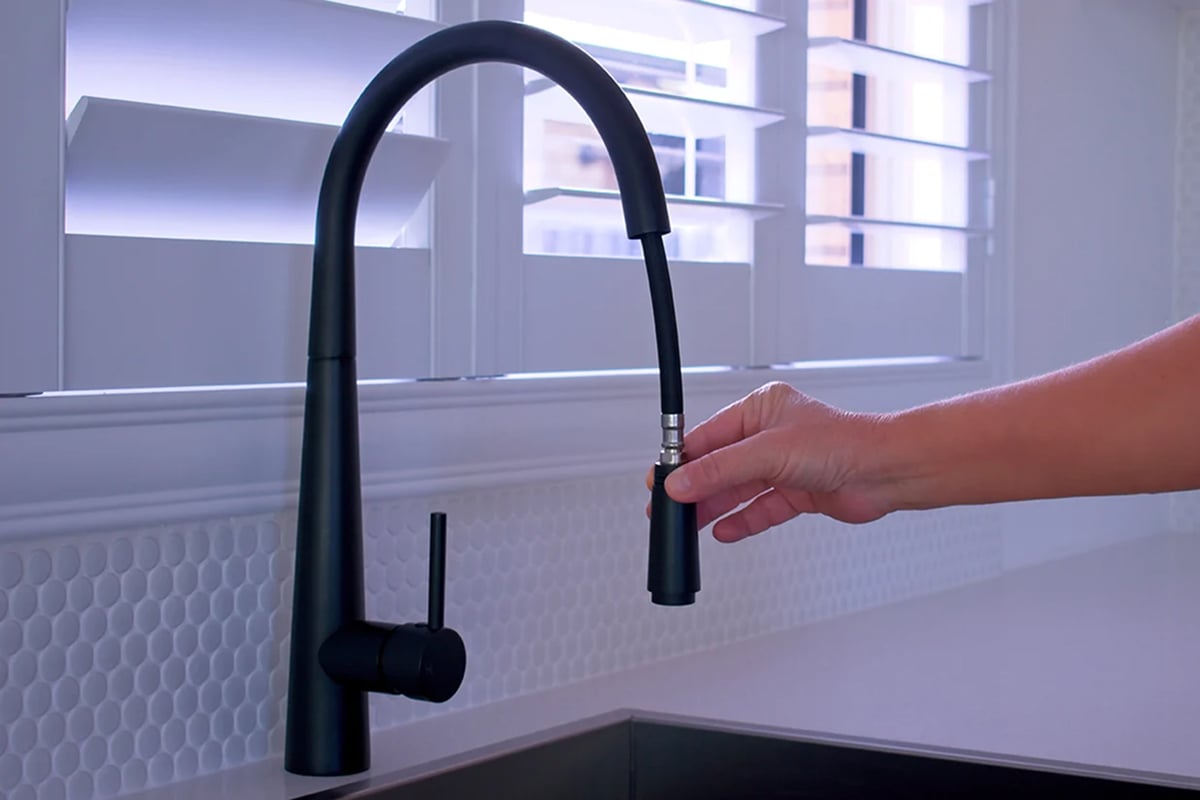 A hand pulls down the spray nozzle of a modern black kitchen faucet—one of the best types of kitchen faucets—over a sink, with white tile backsplash and window shutters in the background. A hand pulls down the spray nozzle of a modern black kitchen faucet—one of the best types of kitchen faucets—over a sink, with white tile backsplash and window shutters in the background.