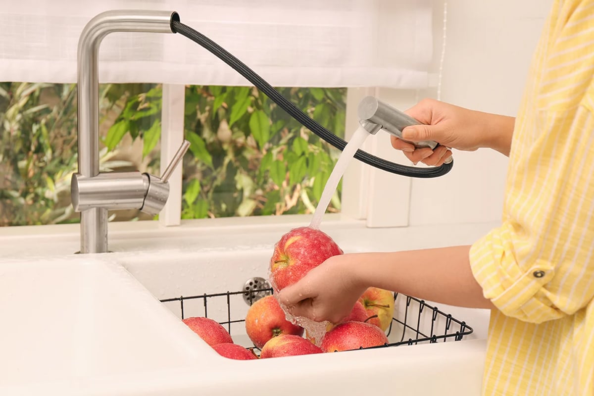 A person in a yellow shirt uses one of the best kitchen faucets—a pull-down model—to wash red apples in a sink, holding one apple under running water, with more apples in a black wire basket below. A person in a yellow shirt uses one of the best kitchen faucets—a pull-down model—to wash red apples in a sink, holding one apple under running water, with more apples in a black wire basket below.