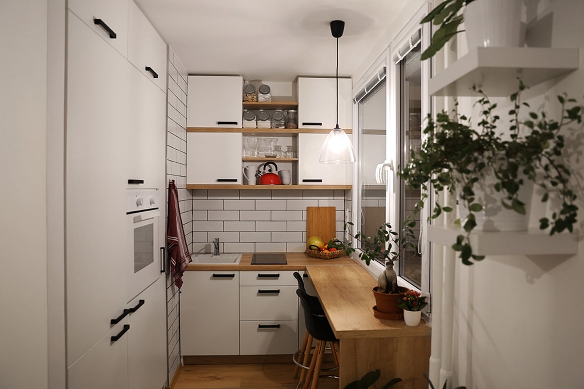 A small kitchen featuring white cabinets, black handles, wood countertops, and open shelves with dishes. Space-saving solutions include a breakfast bar beneath a pendant light, while potted plants and a red kettle add warmth to this modern space.