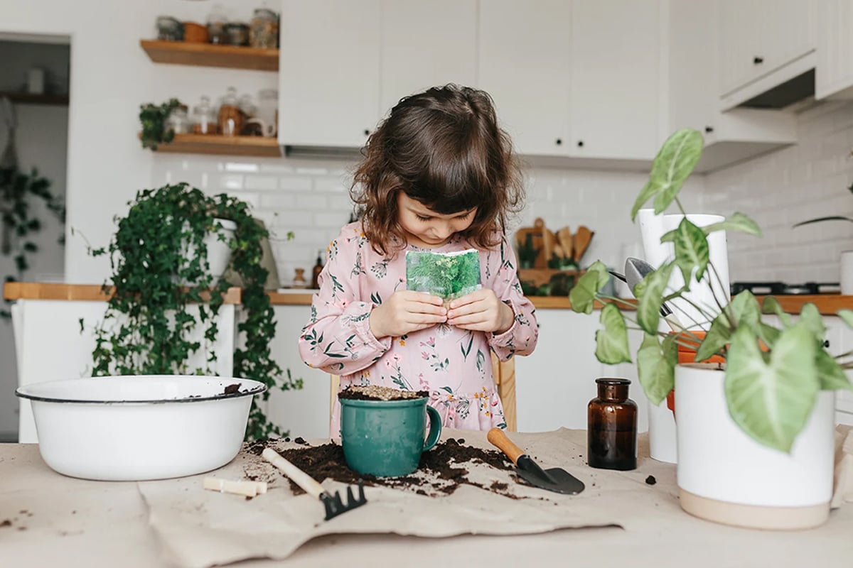 A young girl in a pink floral dress smiles while holding a small green plant over a table covered in soil, gardening tools, and potted plants, enjoying kitchen gardening in a bright indoor garden space.