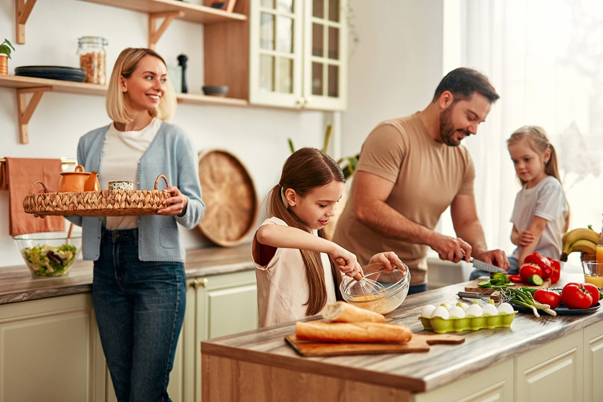 A smiling family prepares food together in a bright kitchen, enjoying the best features perfect for busy families. A woman holds a tray with cups, while fresh produce, bread, and eggs sit on the counter as everyone helps out.