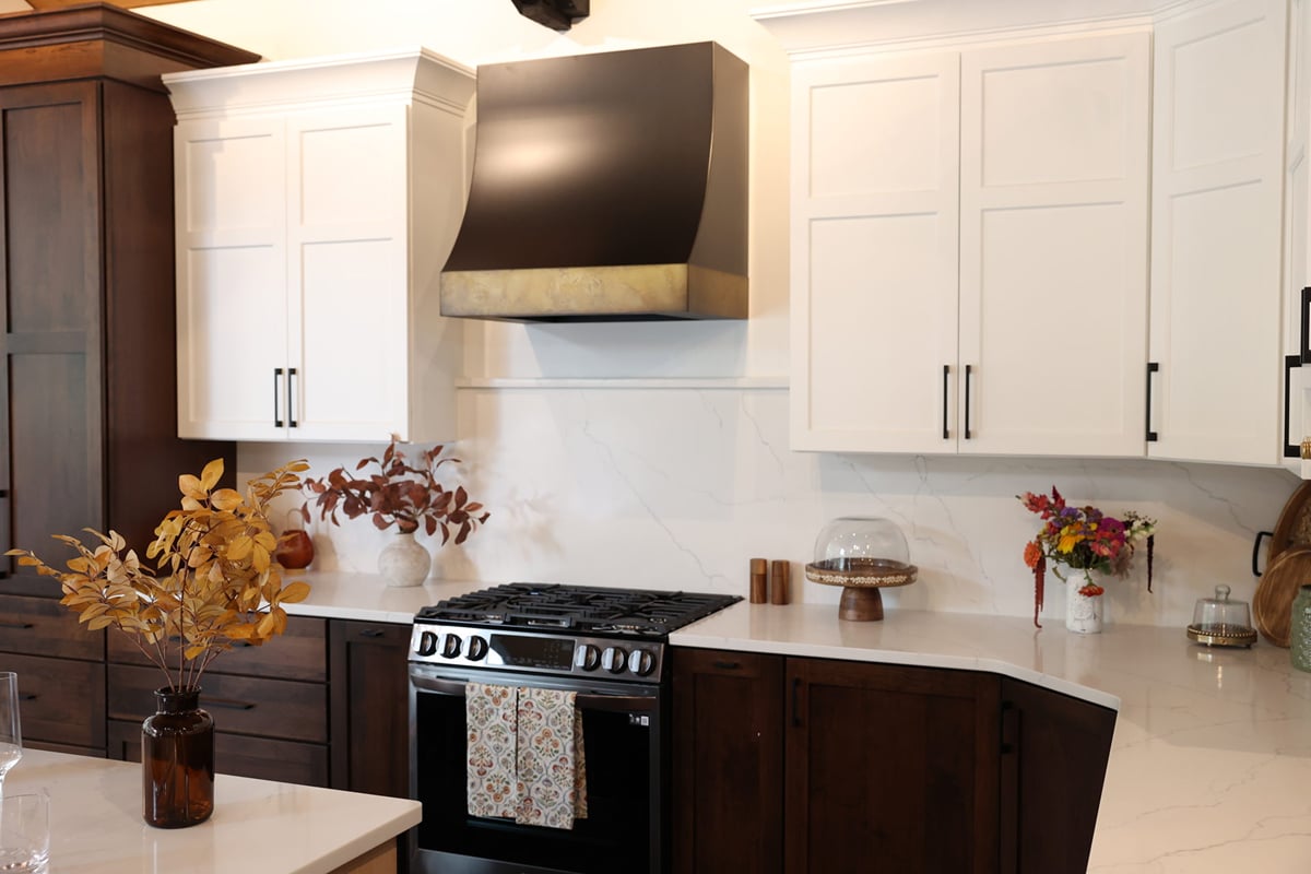 A modern kitchen design features white upper cabinets, dark lower cabinets, a black and gold range hood, a gas stove with a decorative towel, and autumn-themed floral arrangements on the counter, reflecting popular kitchen trends for October 2023.