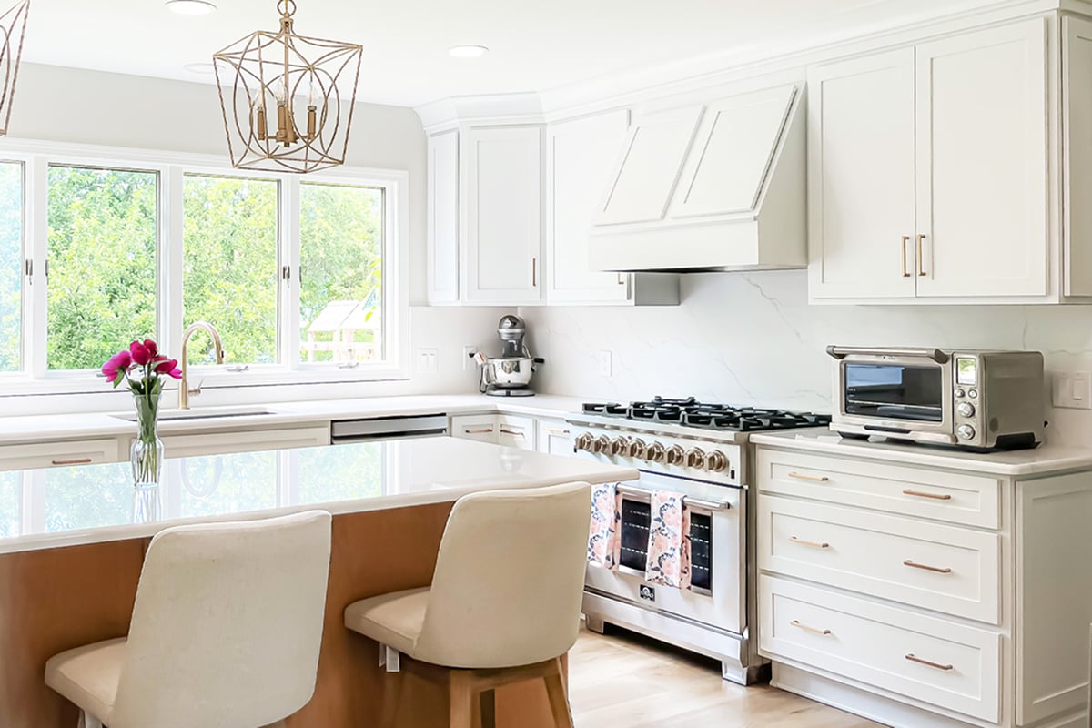 Bright, modern kitchen renovation featuring white cabinets, a large island with two chairs, stainless steel appliances, a vase of pink flowers, and large windows filling the space with natural light.