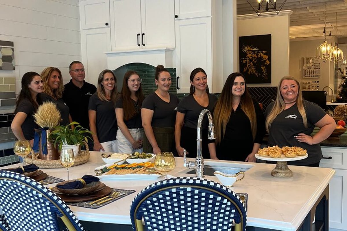 A group of nine adults stands and smiles in a modern Rochester NY kitchen by Kitchens by Oaks, behind a marble island with food and drinks, decorated with plants and elegant table settings.