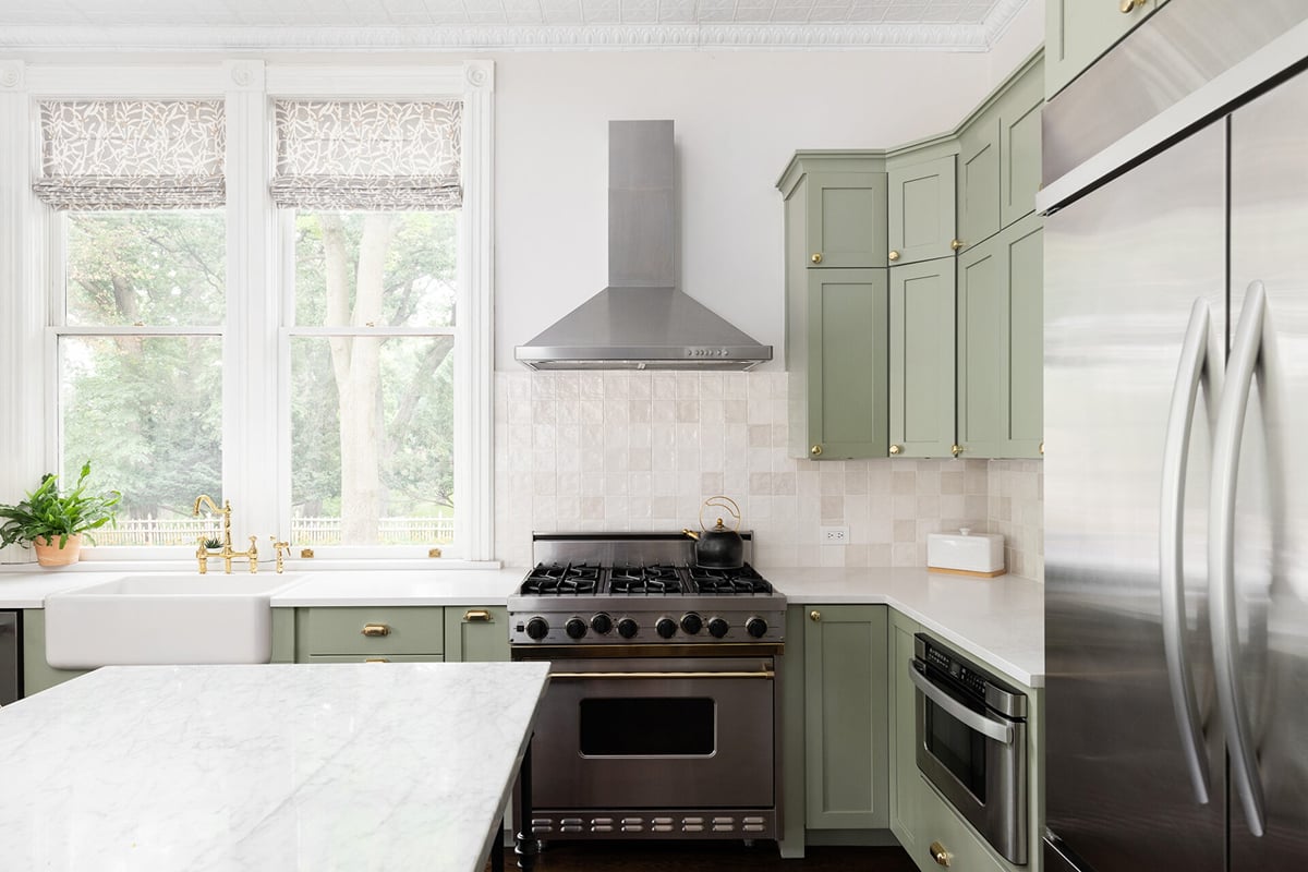 A bright kitchen design with green cabinets, a stainless steel stove and fridge, a marble island, a farmhouse sink, gold fixtures, and two large windows letting in natural light.