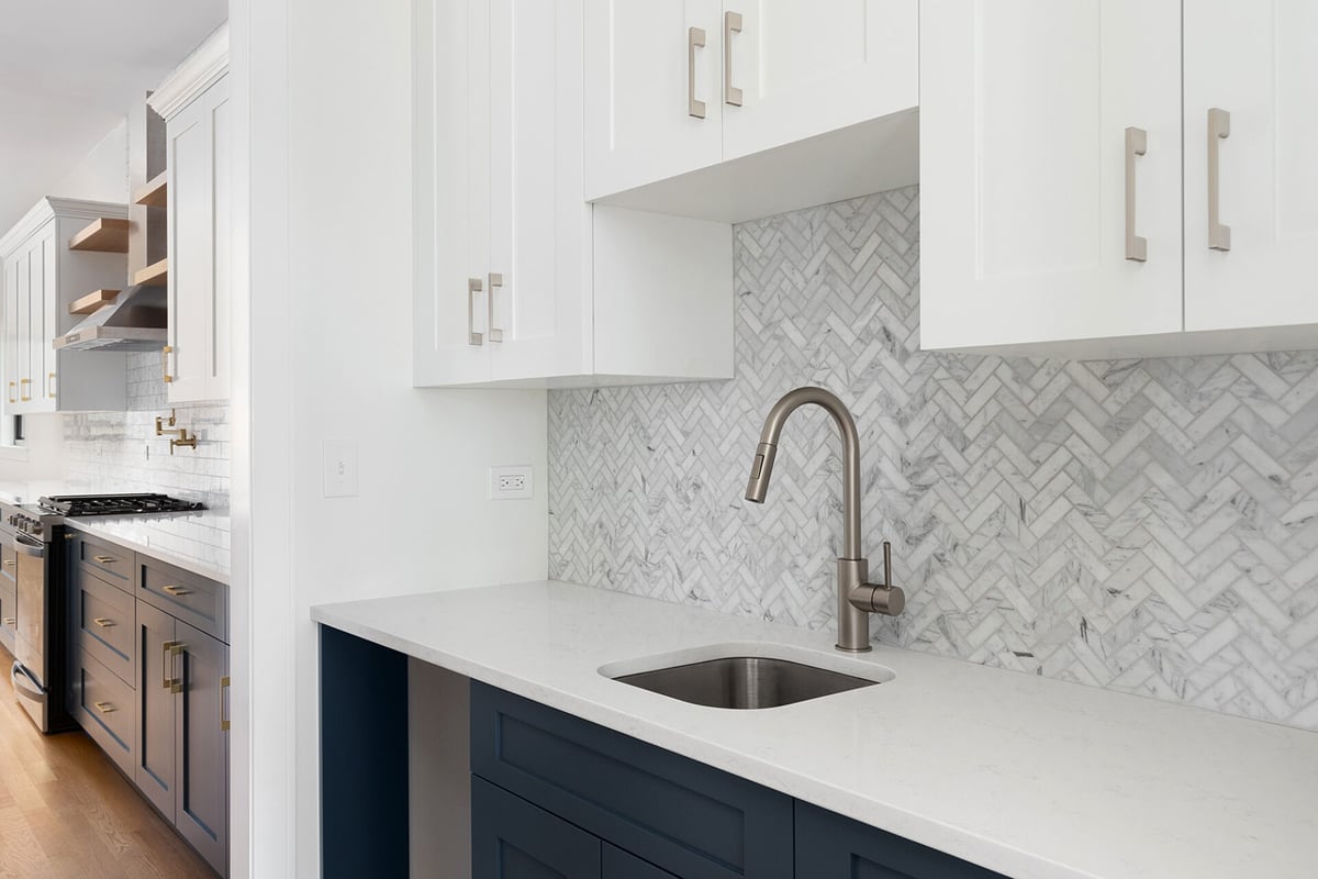 Modern kitchen with white upper cabinets, dark blue lower cabinets, a stainless steel sink, and a herringbone stone tile kitchen backsplash. The background features a stove and open shelves.