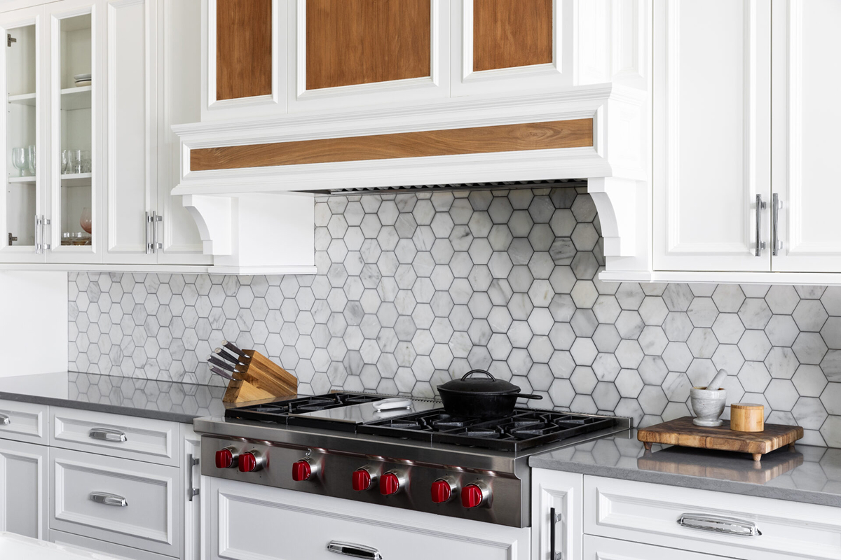 Modern kitchen with white cabinets, gray stone countertops, a hexagonal tile kitchen backsplash, stainless steel gas stove with red knobs, a black pot, knife block, and a small wooden cutting board with kitchen items. Modern kitchen with white cabinets, gray stone countertops, a hexagonal tile kitchen backsplash, stainless steel gas stove with red knobs, a black pot, knife block, and a small wooden cutting board with kitchen items.