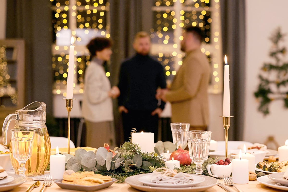 A festively decorated dinner table with candles, food, and glasses in the foreground sets the scene for stress-free entertaining, while three people stand and talk in the softly lit background with string lights in the windows. A festively decorated dinner table with candles, food, and glasses in the foreground sets the scene for stress-free entertaining, while three people stand and talk in the softly lit background with string lights in the windows.