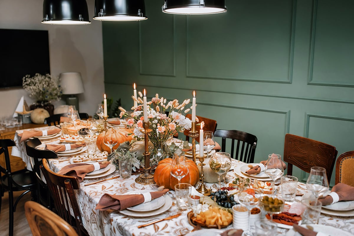 A dining table set for a festive meal with candles, pumpkins, flowers, plates, glasses, and assorted food creates the perfect scene for stress-free entertaining, surrounded by chairs against a green wall under hanging lights.
