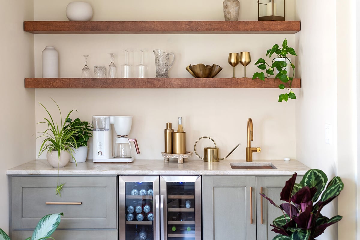 A modern home bar with gray cabinets, a marble countertop, mini fridge, gold faucet, coffee maker, brass bar tools, plants, and open wood shelves holding glasses and decor creates a stylish dry bar against a light wall.