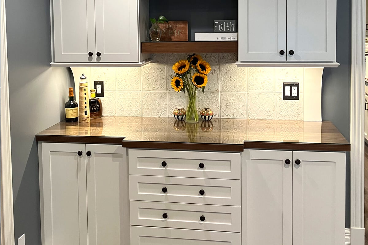 A kitchen counter with white cabinets, a dark wood countertop, a vase of sunflowers, bottles, and decorative items creates a charming home bar. A patterned white backsplash and a small wooden shelf with decor sit above the counter. A kitchen counter with white cabinets, a dark wood countertop, a vase of sunflowers, bottles, and decorative items creates a charming home bar. A patterned white backsplash and a small wooden shelf with decor sit above the counter.