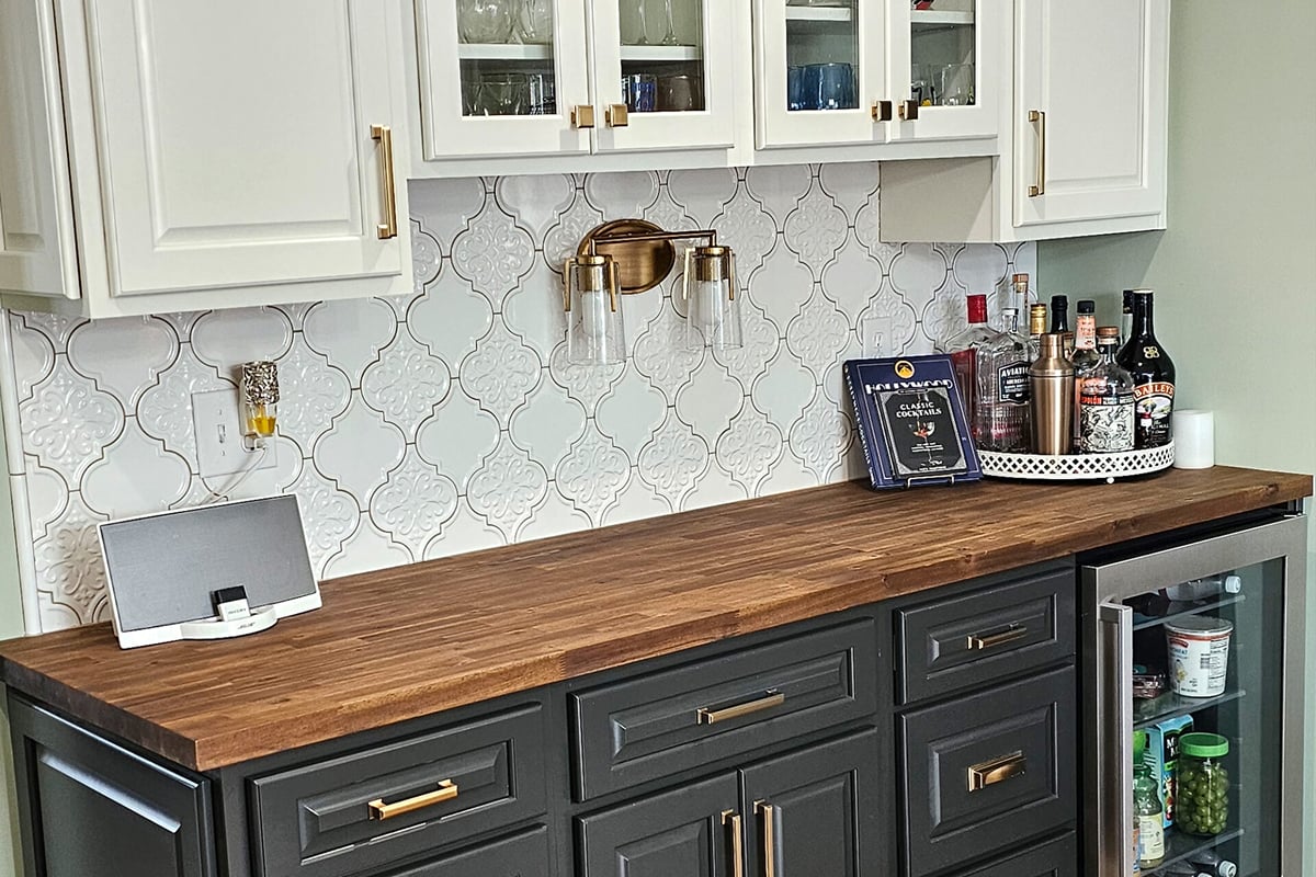 A stylish home bar setup features a wood kitchen counter, dark lower cabinets with gold handles, white glass-front uppers, and a glass-front mini fridge. Bottles, a tray, a cookbook, and a speaker complete this elegant dry bar space. A stylish home bar setup features a wood kitchen counter, dark lower cabinets with gold handles, white glass-front uppers, and a glass-front mini fridge. Bottles, a tray, a cookbook, and a speaker complete this elegant dry bar space.