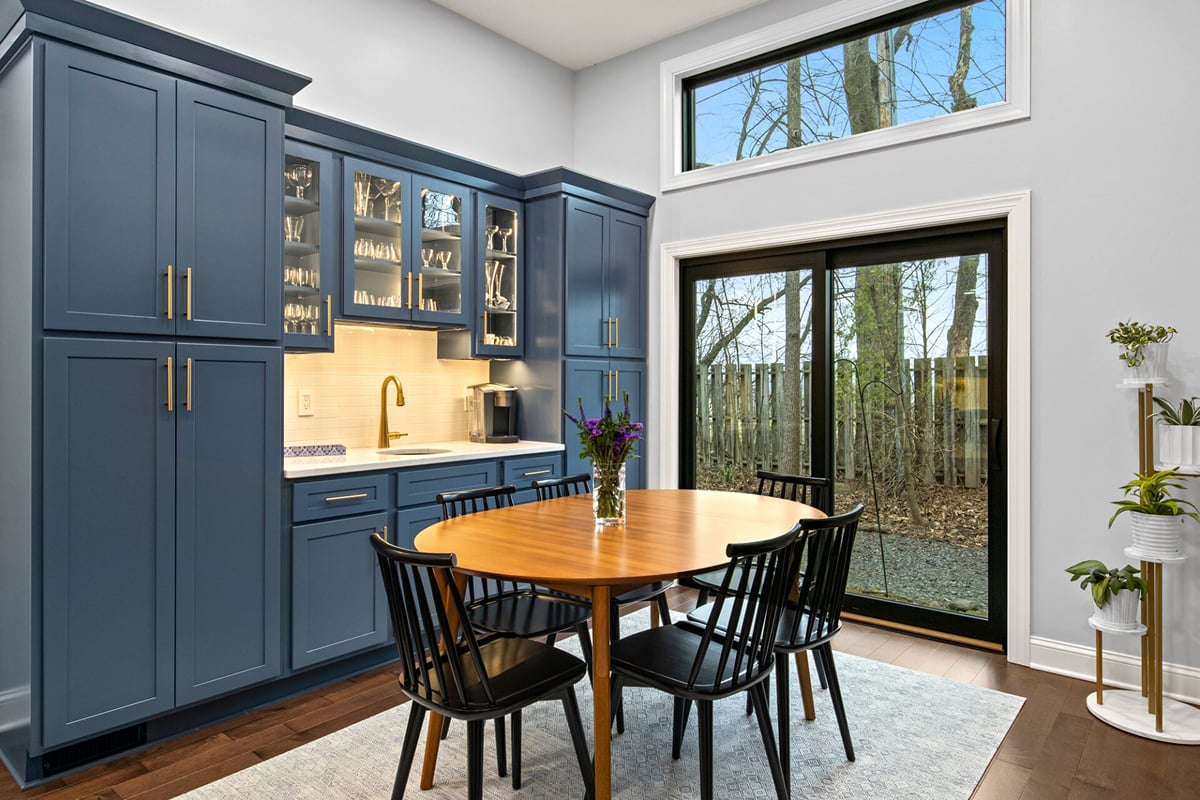 A modern kitchen with blue cabinets, a stylish wet bar, a round wooden table with four black chairs, under-cabinet lighting, and large windows framing trees outside. A vase of flowers sits on the table, with plants on a white stand nearby. A modern kitchen with blue cabinets, a stylish wet bar, a round wooden table with four black chairs, under-cabinet lighting, and large windows framing trees outside. A vase of flowers sits on the table, with plants on a white stand nearby.