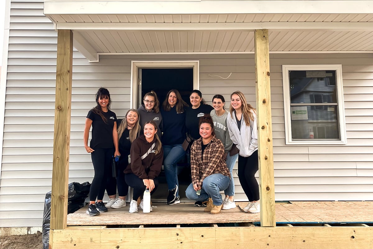 Nine women pose and smile together on the porch of a house under construction, building community with Habitat for Humanity. All are casually dressed and appear happy, enjoying their time volunteering among wooden posts and unfinished flooring.