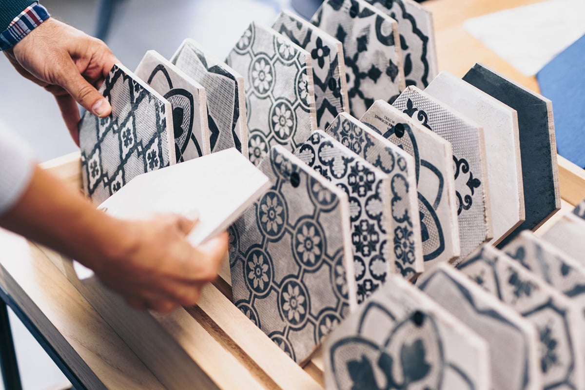 A person selects from a wooden display rack holding various black-and-white patterned ceramic tiles, featuring geometric and floral designs—perfect choices for stylish waterproof flooring. A person selects from a wooden display rack holding various black-and-white patterned ceramic tiles, featuring geometric and floral designs—perfect choices for stylish waterproof flooring.