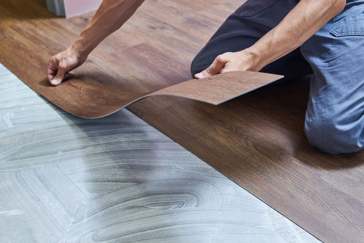 A person installs brown waterproof flooring, carefully placing a plank over adhesive on the subfloor, while wearing blue pants and kneeling on the ground. A person installs brown waterproof flooring, carefully placing a plank over adhesive on the subfloor, while wearing blue pants and kneeling on the ground.