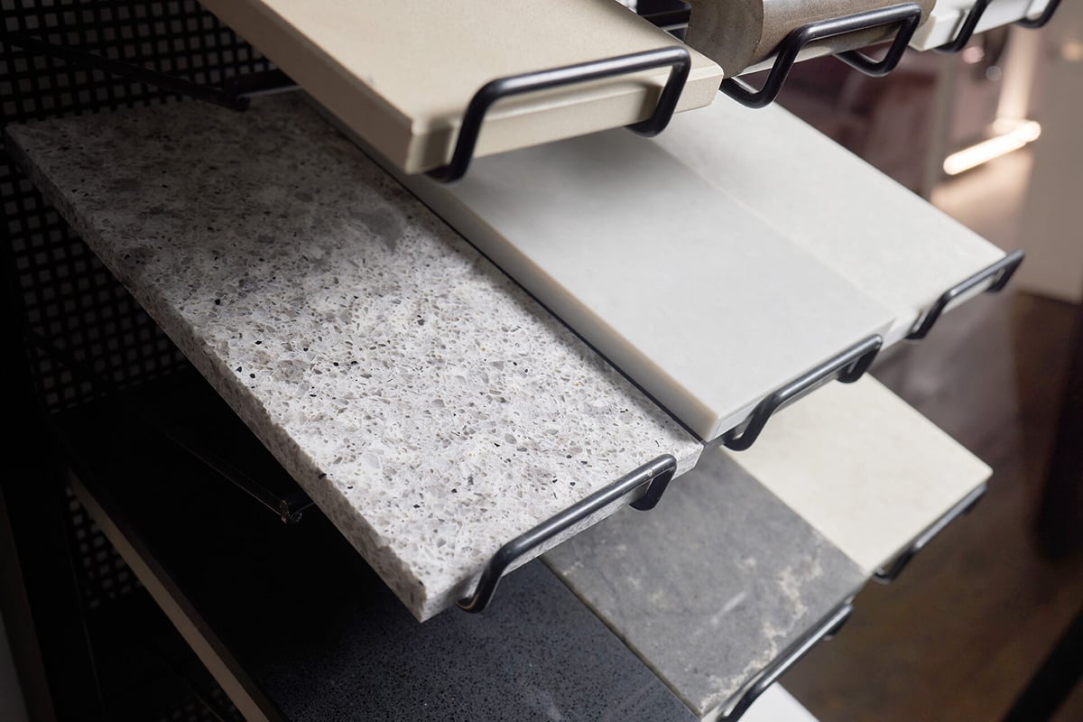 Close-up of several rectangular countertop samples in different colors and textures, displayed on a black metal rack. The surfaces, ideal for pairing with waterproof flooring, include speckled, solid, and marbled patterns in gray and beige shades.