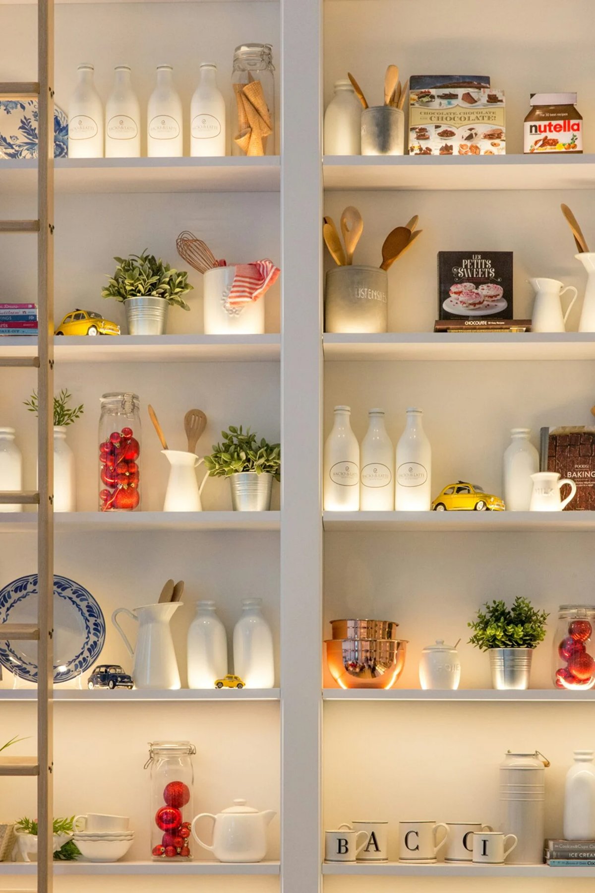 A well-lit kitchen shelf from a recent kitchen remodel displays white bottles, mugs, jars with cherries, small plants, yellow toy cars, cookbooks, utensils, and decorative items including a Nutella jar and blue-patterned plates.