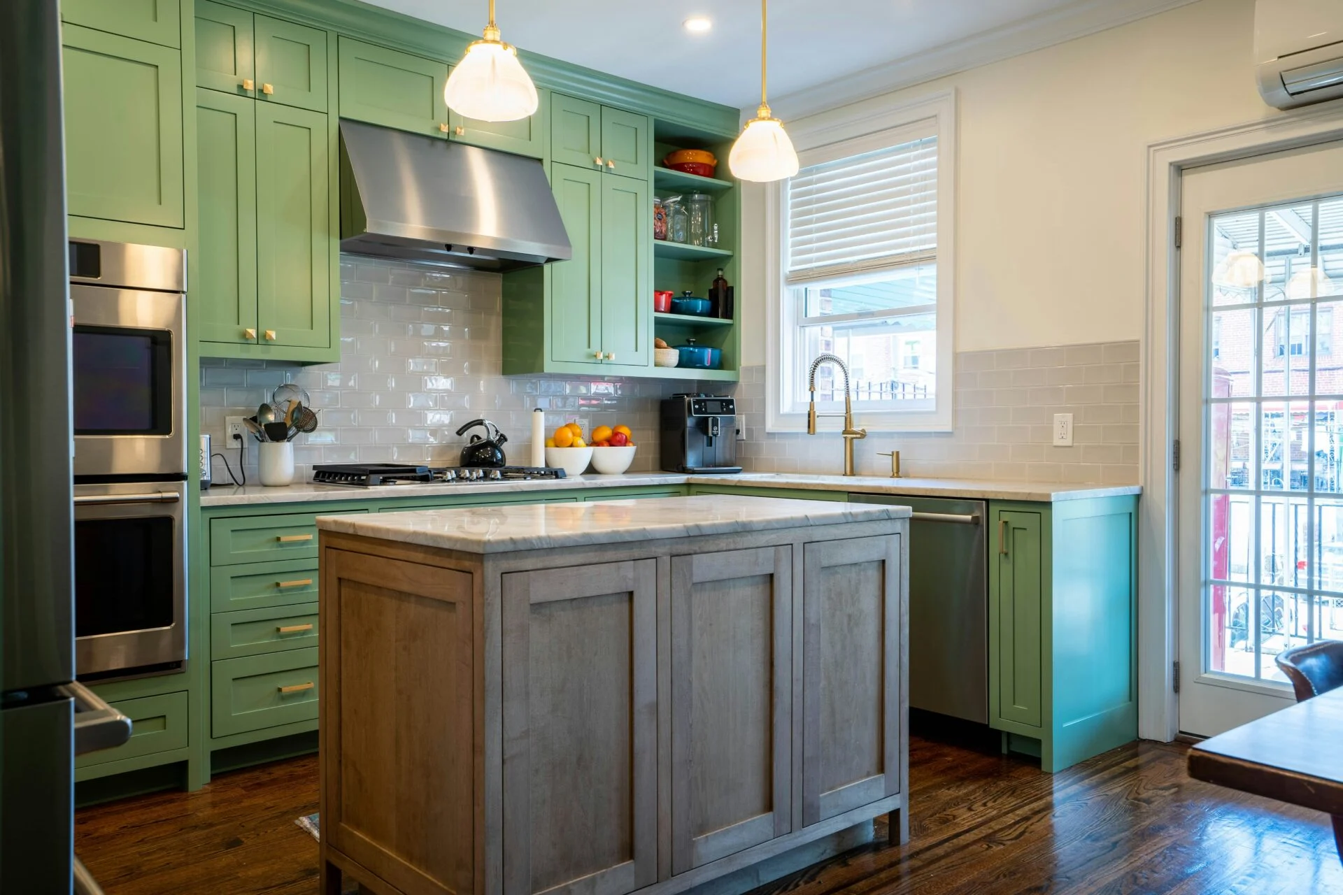 A modern kitchen remodel with green cabinets, stainless steel appliances, a marble island, pendant lights, wooden floors, and a window and glass door welcoming natural light. Bowls of fruit and kitchen items sit on the counters.