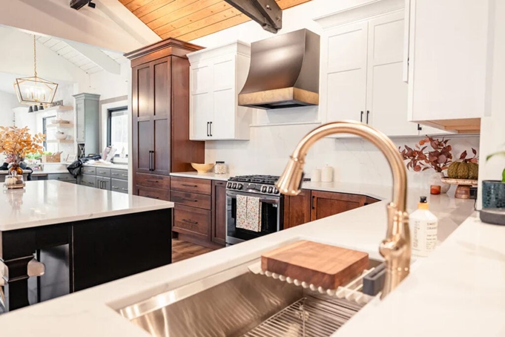Modern kitchen remodel with white and wood cabinetry, stainless steel oven, large island featuring a built-in sink, gold faucet, soap dispenser, cutting board, and decorative plants on the counter.