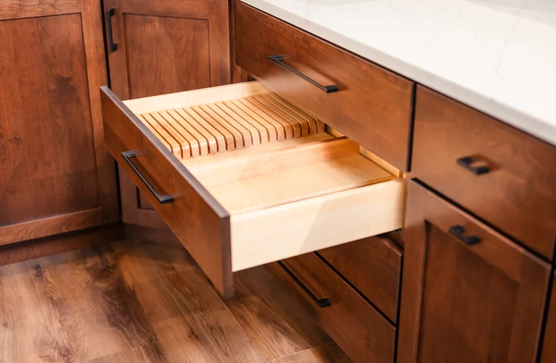A wooden kitchen drawer with built-in knife slots is open, displaying organized spaces for knives—an elegant touch to any kitchen remodel, set in cabinetry with dark handles and a white countertop above.