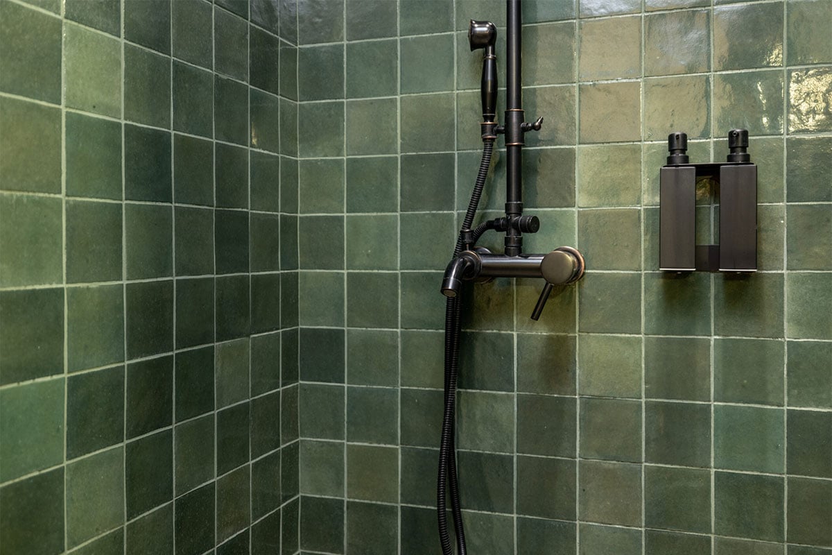 Close-up of a shower with green tiled walls, a black showerhead and handle, and two wall-mounted black soap dispensers—perfect inspiration for your next bathroom or kitchen remodel.