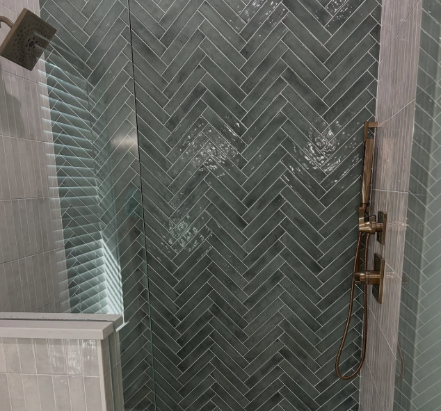 A modern shower with glossy dark green herringbone tile, a rainfall showerhead on the left, and a vertical bar with a handheld shower and controls on the right—perfect inspiration for your next bath or kitchen remodel. A glass enclosure corner is visible. A modern shower with glossy dark green herringbone tile, a rainfall showerhead on the left, and a vertical bar with a handheld shower and controls on the right—perfect inspiration for your next bath or kitchen remodel. A glass enclosure corner is visible.