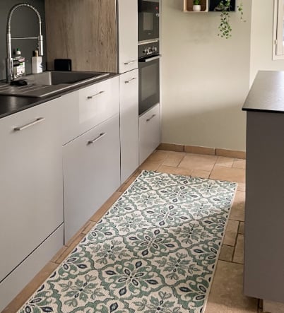 A modern kitchen remodel with gray cabinets, a stainless steel sink, built-in appliances, beige tile floor, and a long patterned rug in green and blue floral motifs. A small plant hangs on the wall.