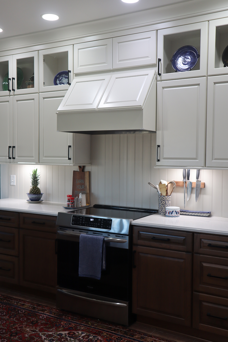 A modern kitchen remodel features white upper cabinets, dark lower cabinets, a stainless steel stove with a towel hanging on the handle, a knife rack, utensils, a small potted plant, and decorative plates on display.