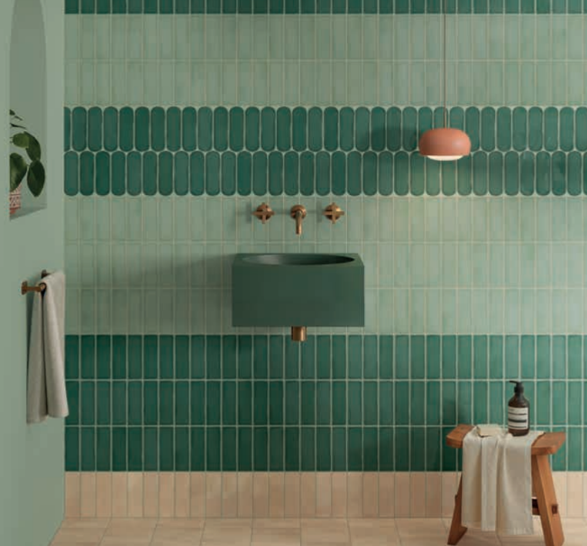 A stylish bathroom reminiscent of a luxe kitchen remodel, with green vertical tiles, a dark green sink, wall-mounted brass faucet, coral wall light, towel on a hook, small wooden stool with accessories, and a leafy plant partially visible on the left. A stylish bathroom reminiscent of a luxe kitchen remodel, with green vertical tiles, a dark green sink, wall-mounted brass faucet, coral wall light, towel on a hook, small wooden stool with accessories, and a leafy plant partially visible on the left.