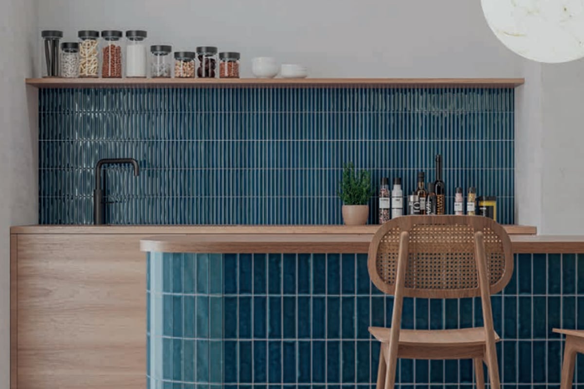 A modern kitchen remodel featuring a blue tiled backsplash and counter, wooden cabinets, a wooden chair with a woven back, black faucet, assorted jars on a shelf, bottles, and a small potted plant.