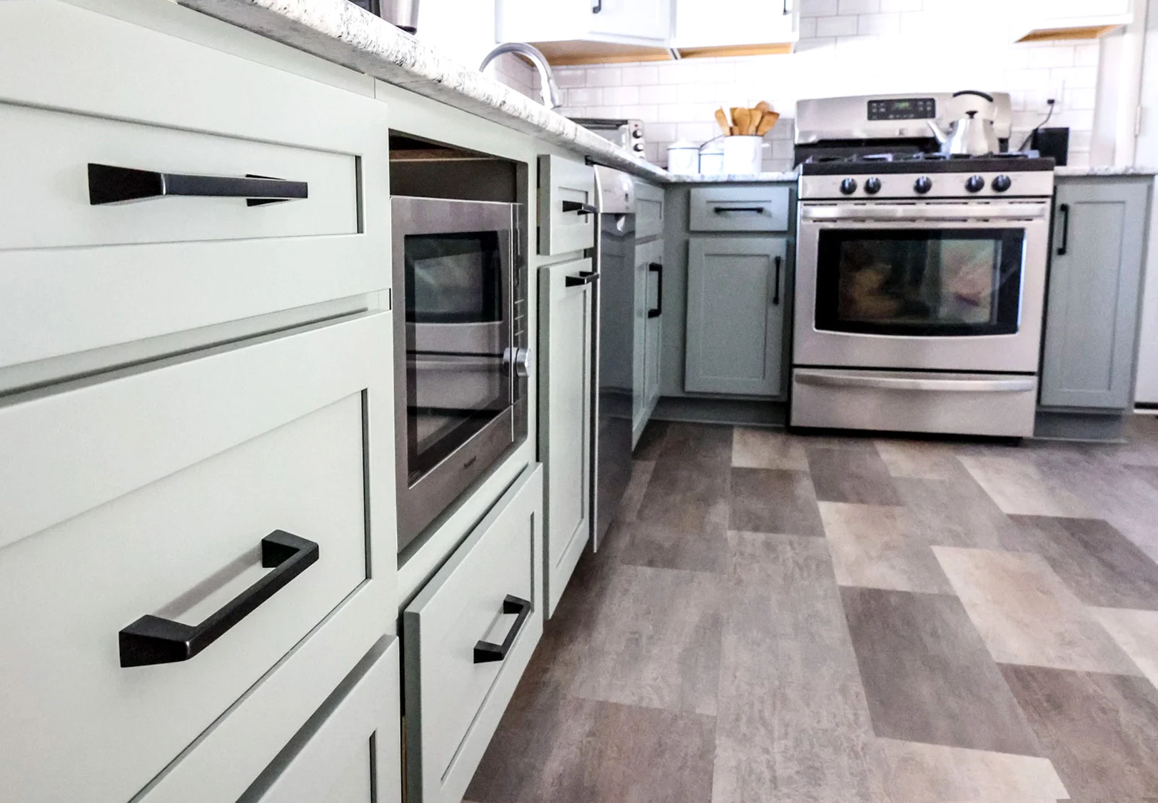 Modern kitchen remodel with light green cabinets, black handles, a built-in microwave, gas stove with oven, and mixed light and dark gray flooring. White countertops display kitchen items in the background for a fresh update. Modern kitchen remodel with light green cabinets, black handles, a built-in microwave, gas stove with oven, and mixed light and dark gray flooring. White countertops display kitchen items in the background for a fresh update.