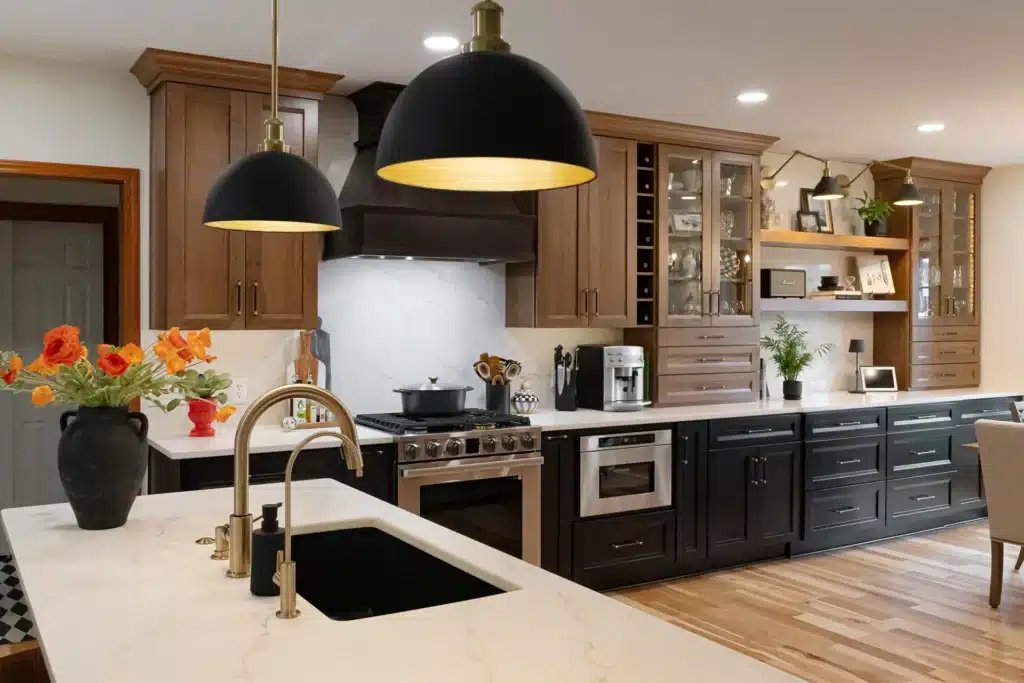 Modern kitchen remodel with a marble island, black and wood cabinets, gold fixtures, black pendant lights, and open shelves decorated with plants and books. A vase with orange flowers sits near the sink.