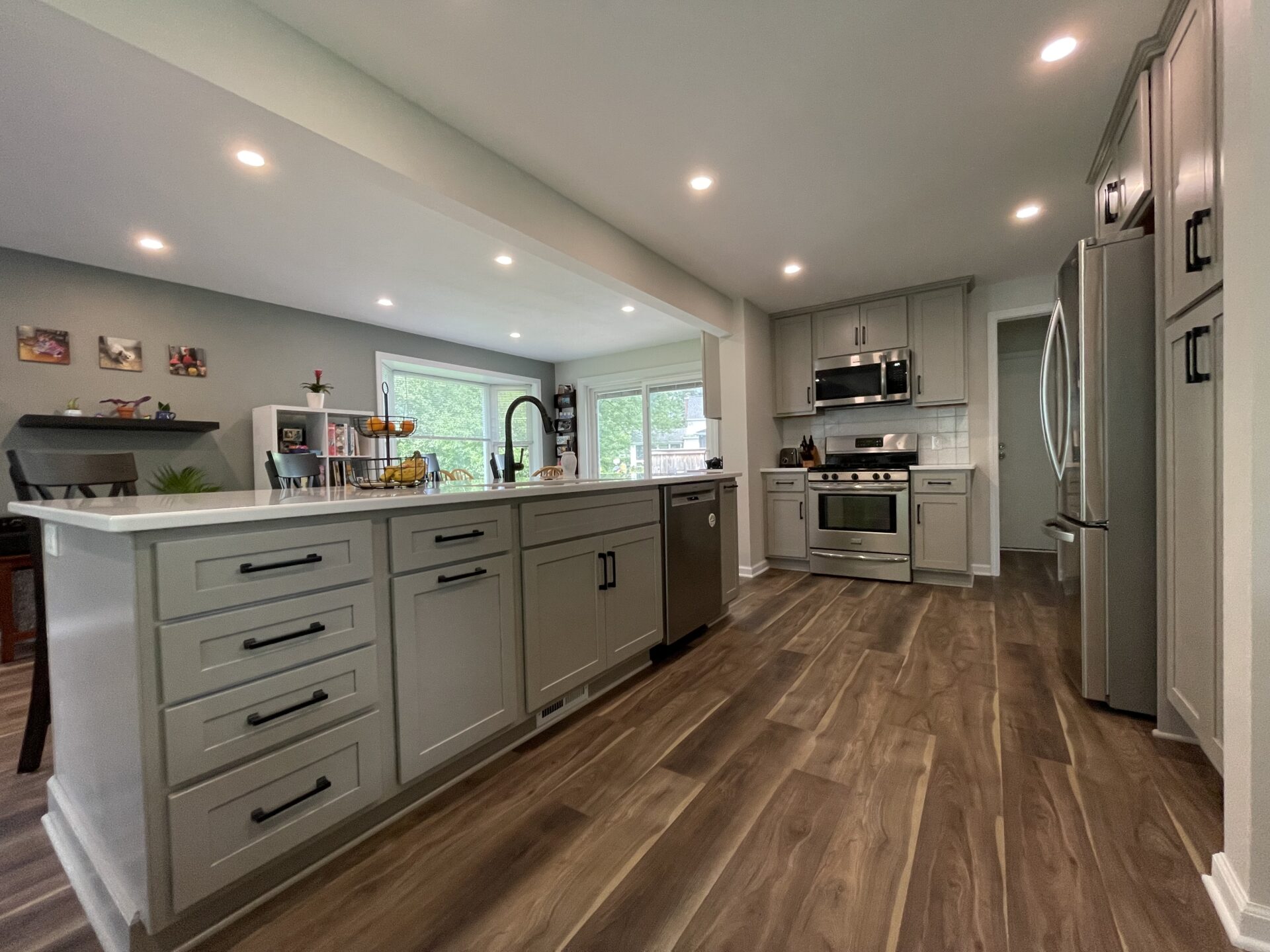 Modern kitchen remodel with gray cabinets, stainless steel appliances, an island with a sink and dishwasher, wood-look flooring, recessed lighting, and large windows providing natural light. Modern kitchen remodel with gray cabinets, stainless steel appliances, an island with a sink and dishwasher, wood-look flooring, recessed lighting, and large windows providing natural light.