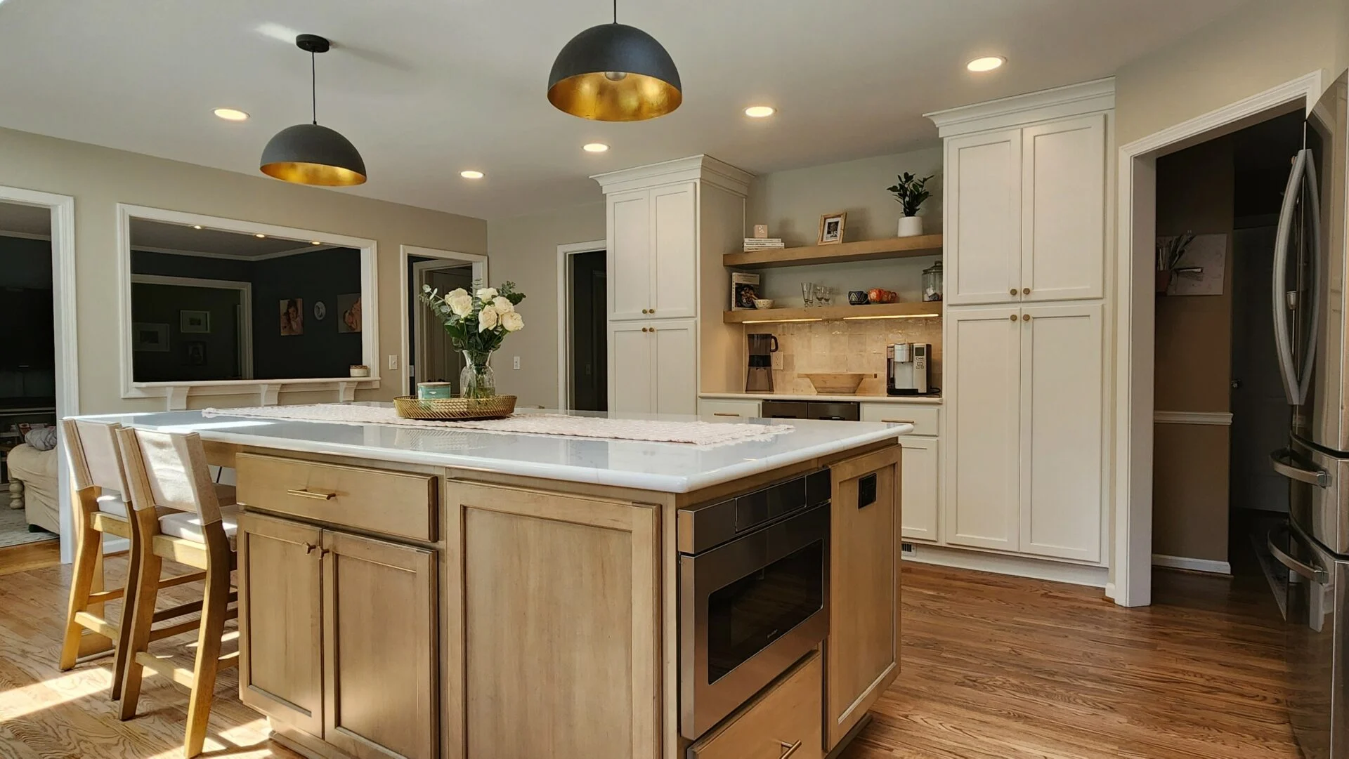 Modern kitchen remodel with light wood floors, white cabinets, a large island with chairs, built-in oven, and pendant lights. A vase of flowers sits on the island, and shelves with decor are visible in the background. Modern kitchen remodel with light wood floors, white cabinets, a large island with chairs, built-in oven, and pendant lights. A vase of flowers sits on the island, and shelves with decor are visible in the background.