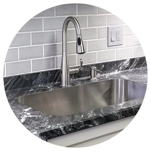 A modern stainless steel sink with a high-arc faucet is set in a dark marble countertop—an ideal centerpiece for your kitchen remodel—complete with a soap dispenser and white subway tile backsplash in the background. A modern stainless steel sink with a high-arc faucet is set in a dark marble countertop—an ideal centerpiece for your kitchen remodel—complete with a soap dispenser and white subway tile backsplash in the background.