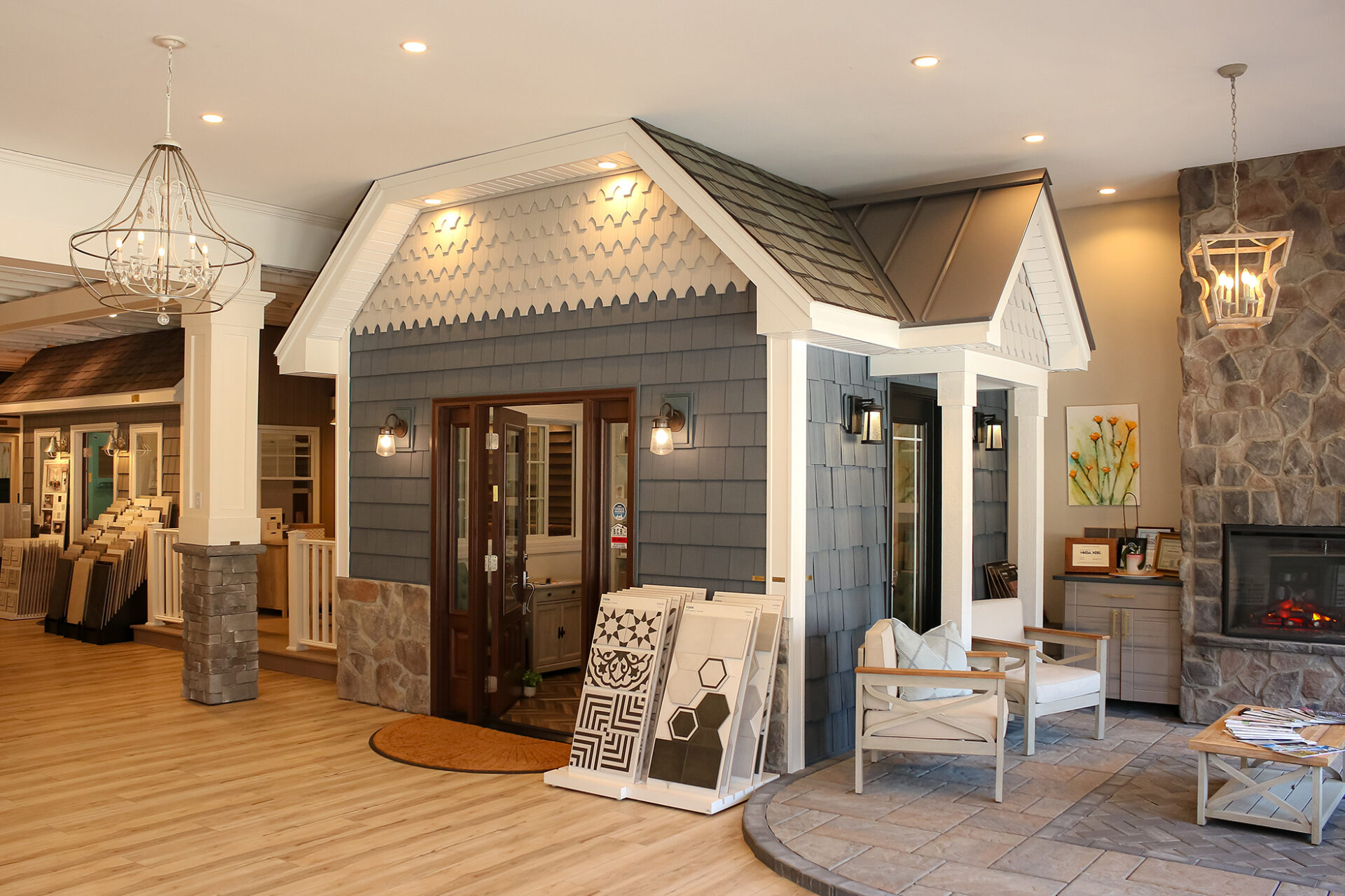 A showroom with a small house display featuring gray siding, a shingled roof, and double doors. Inside, see fireplace, chairs, flooring samples, decorative lighting, and inspiration for your next kitchen remodel in the spacious, well-lit interior. A showroom with a small house display featuring gray siding, a shingled roof, and double doors. Inside, see fireplace, chairs, flooring samples, decorative lighting, and inspiration for your next kitchen remodel in the spacious, well-lit interior.